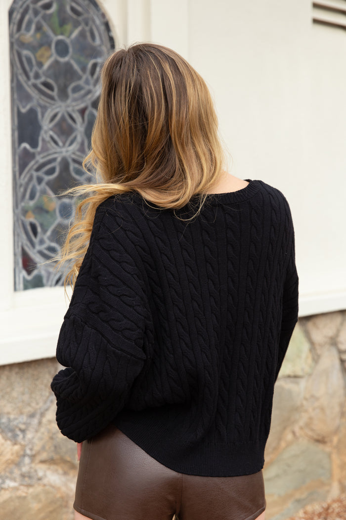 A woman viewed from behind wearing a black cable-knit sweater and brown faux-leather shorts, standing outdoors in front of a white wall and stone base.