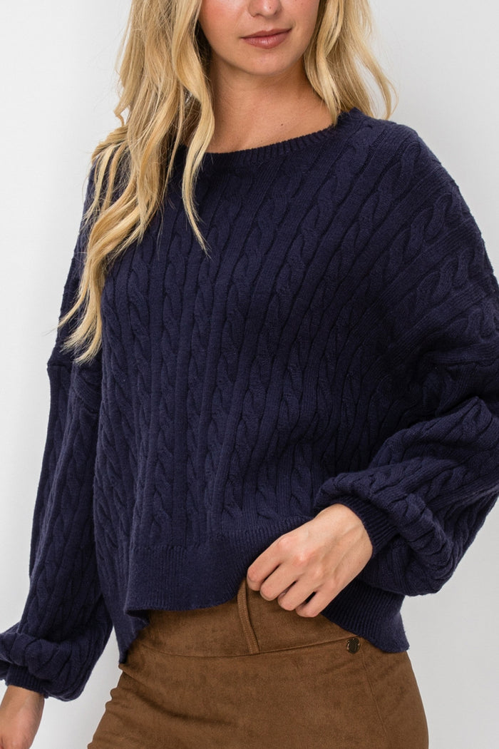 A woman wearing a navy blue cable-knit sweater and a brown mini skirt, photographed against a light gray studio background.