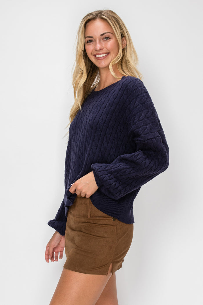 A woman shown from the side wearing a navy blue cable-knit sweater and a brown mini skirt, photographed against a light gray studio background.