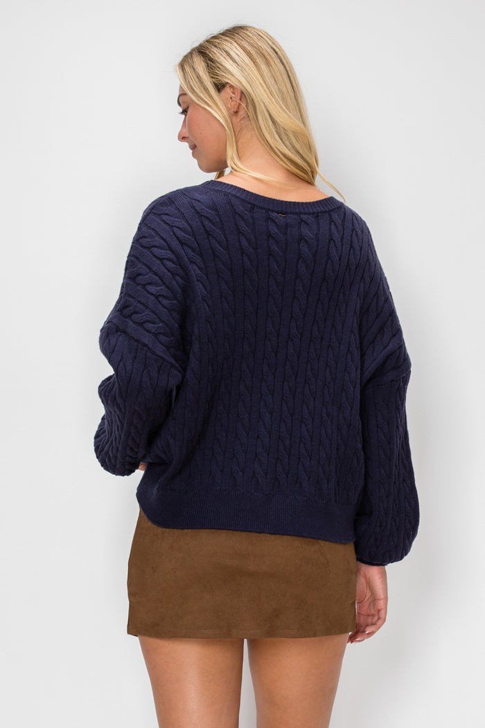 A woman shown from the back wearing a navy blue cable-knit sweater and a brown mini skirt, photographed against a light gray studio background.
