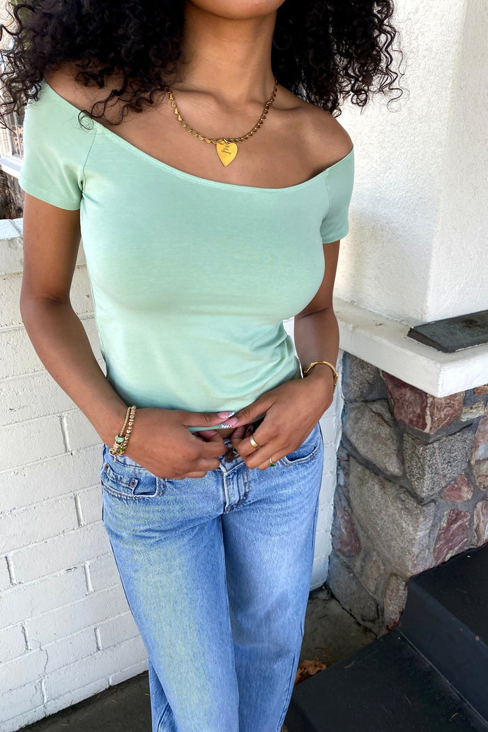 Person wearing a light green off-shoulder top and blue jeans standing against a white brick wall.
