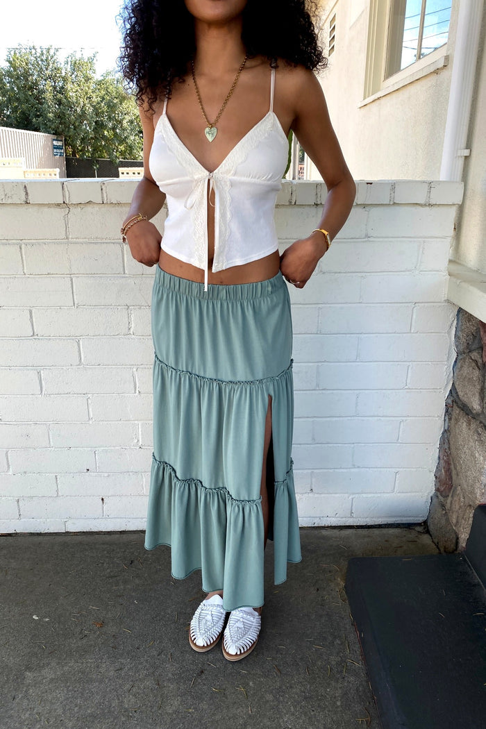 Woman wearing a white top and green skirt standing outdoors against a white brick wall.