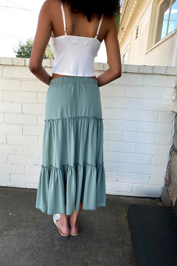 Person wearing a white top and green tiered skirt standing against a white brick wall.