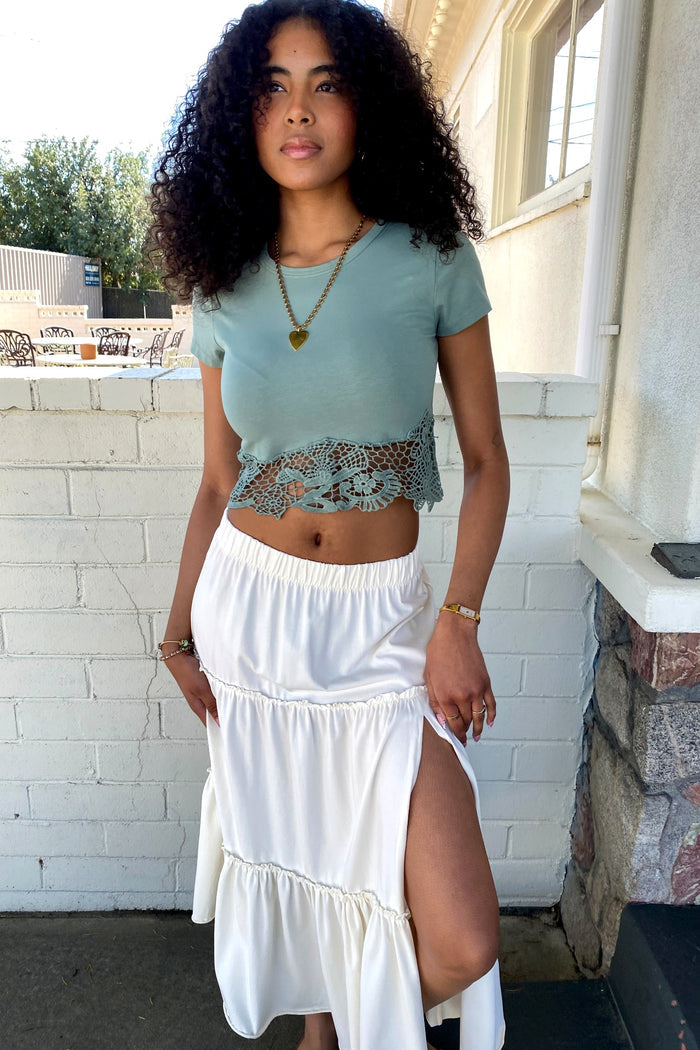 Woman wearing a light blue top with lace detail and a white skirt with a slit, standing against a white brick wall.
