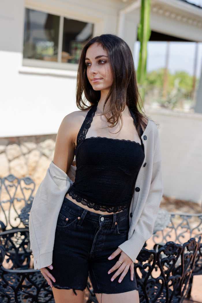 Woman wearing a black top and denim shorts with a beige cardigan outdoors.