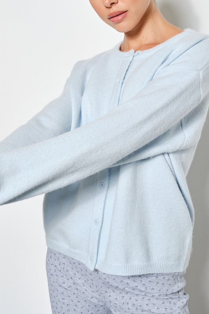 A close-up of a person wearing a light blue knit cardigan with a button front and long sleeves, paired with light patterned pants against a plain light background.