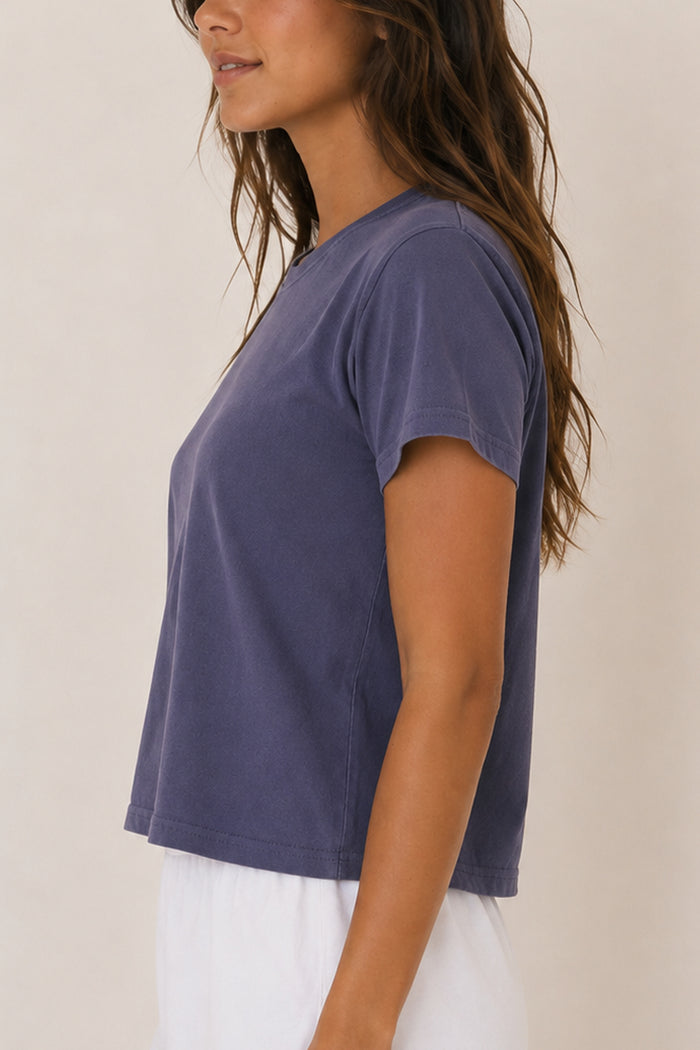 A woman wearing a short-sleeve crewneck t-shirt in a washed slate blue color, paired with white pants, shown from a side profile against a plain beige background.