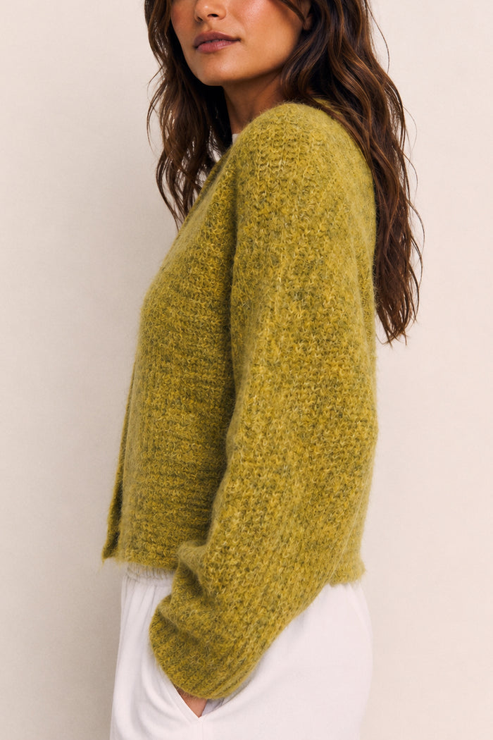 A woman wearing a fuzzy, textured olive green knit cardigan sweater and white pants, shown from a side profile against a plain light beige background.