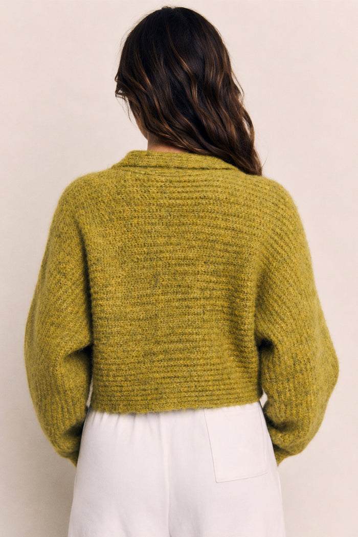 A woman viewed from behind wearing a textured, chunky knit cropped sweater in a muted olive yellow-green, paired with white pants, against a plain beige background.