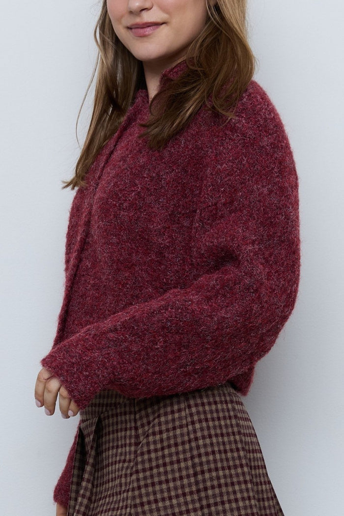 A woman wearing a fuzzy, textured knit cardigan in a deep burgundy red, paired with a brown and beige gingham skirt, photographed against a plain light gray background.