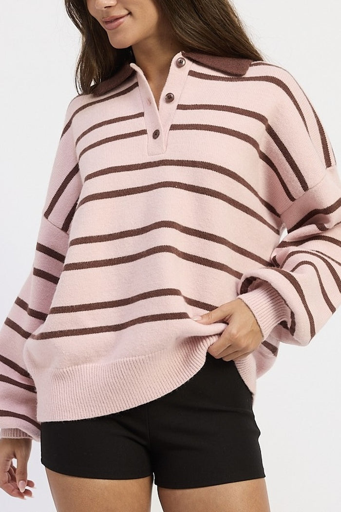 A woman wearing a pink and brown horizontally striped knit polo sweater with a buttoned collar, paired with black shorts, against a plain light gray background.