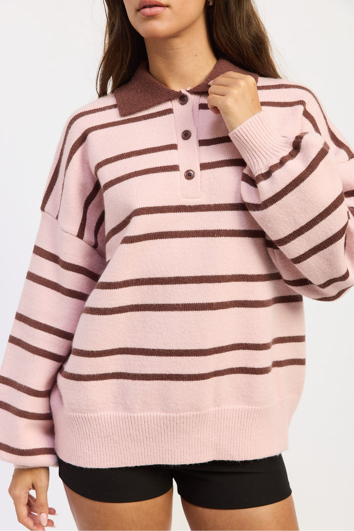 A woman wearing a pink and brown horizontally striped knit polo sweater with a three-button placket, paired with black shorts, against a plain white background.
