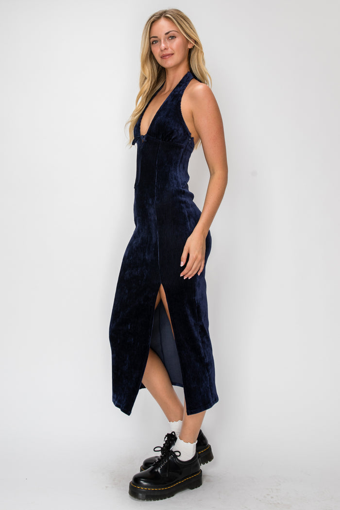 A woman modeling a sleeveless navy blue velvet midi dress with a plunging neckline and a high side slit, paired with black leather platform shoes, against a plain white studio background.