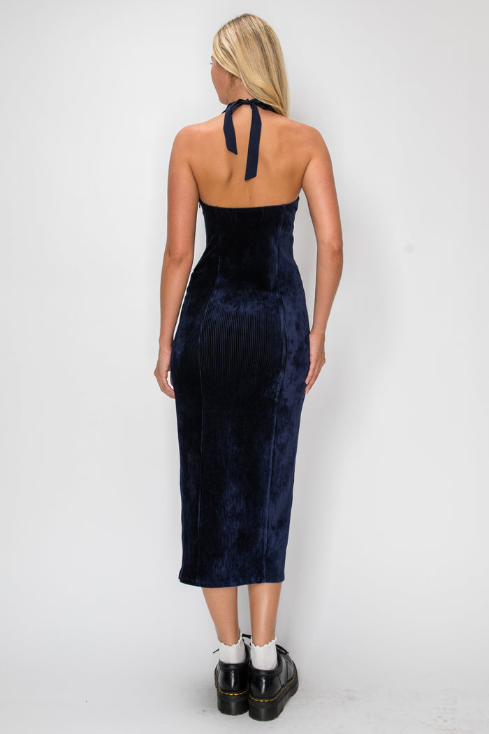 A woman viewed from the back wearing a dark navy velvet halter midi dress with a tie at the neck, paired with white socks and black leather shoes, against a plain light gray studio background.