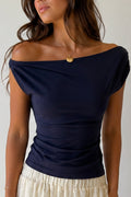 A woman wearing a navy blue off-the-shoulder top with ruched detailing at the waist, paired with a cream-colored textured skirt. She is wearing a delicate gold necklace.