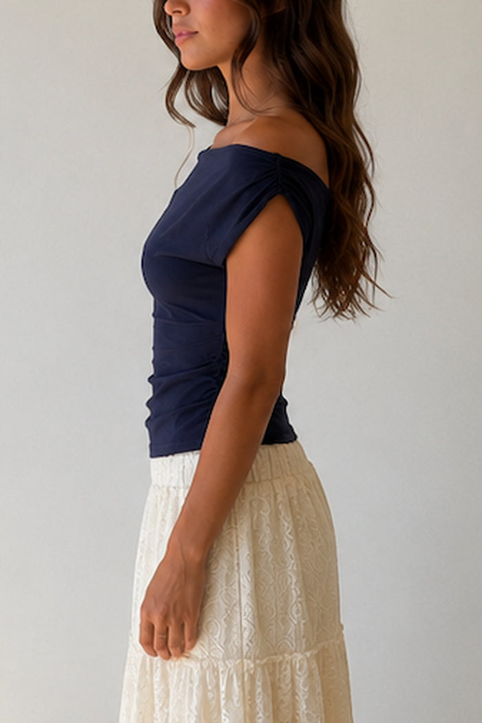 A woman wearing a navy blue off-the-shoulder top with ruched detailing at the waist, paired with a cream-colored textured skirt. She is wearing a delicate gold necklace.