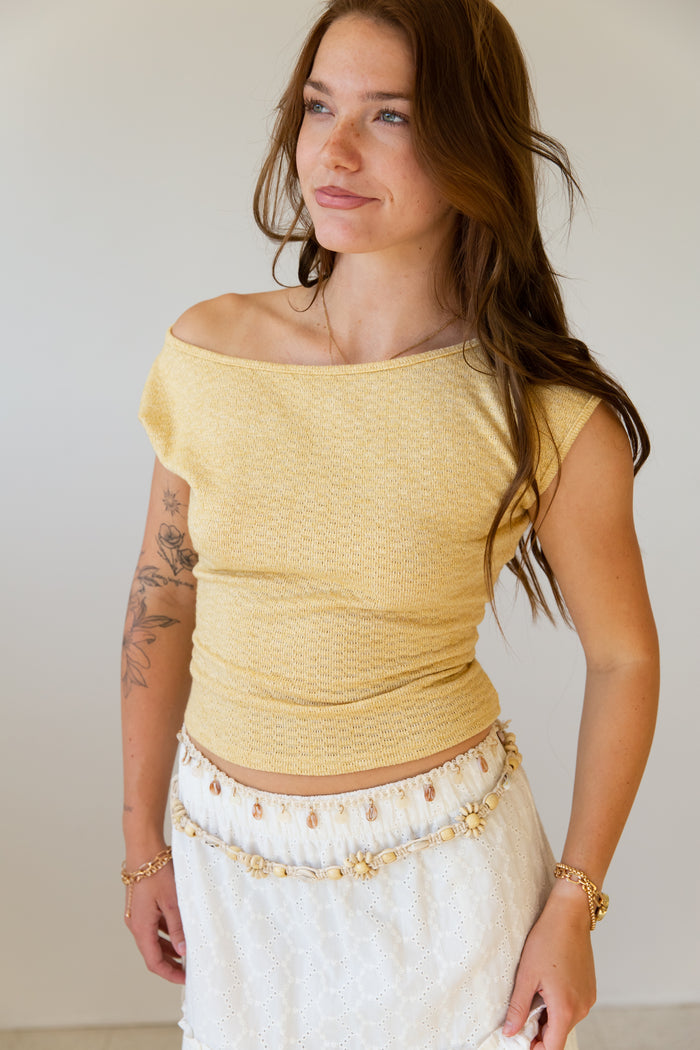 Woman wearing a yellow off-shoulder top and white pants with gold accents.