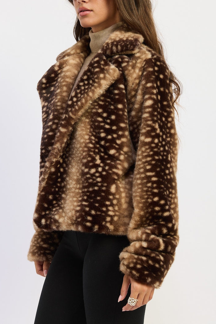 A woman wearing a brown faux fur jacket with a cream spotted deer-print pattern, paired with a beige turtleneck and black pants, shown from the chest down against a plain white background.