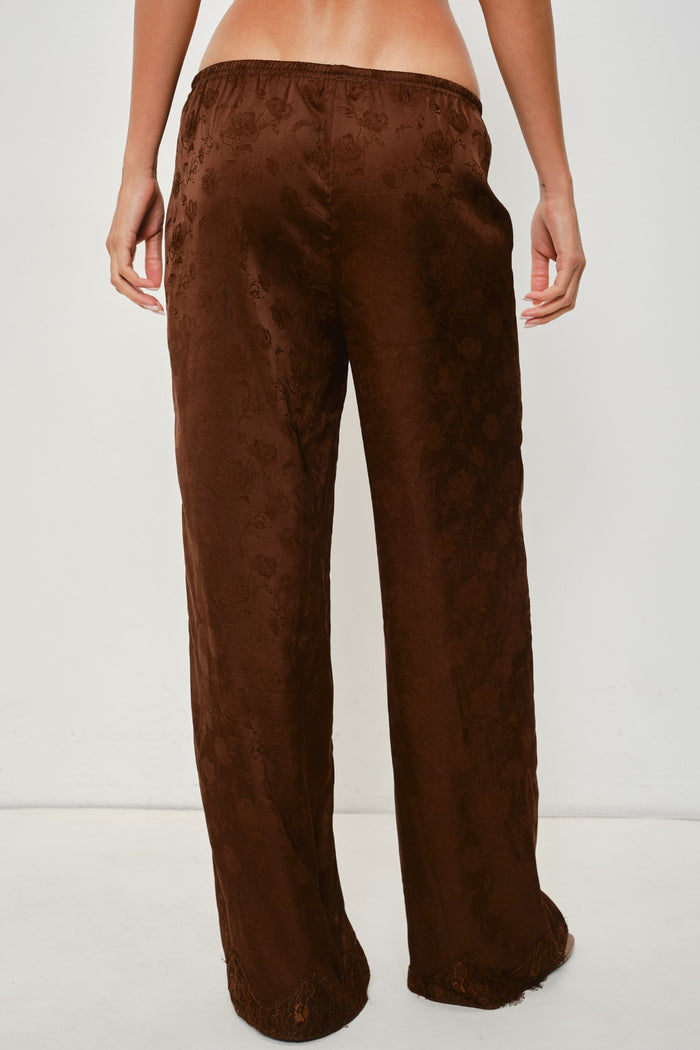 A person shown from the back wearing loose, wide-leg brown pants with a subtle tonal floral jacquard pattern, photographed against a plain light gray studio background.