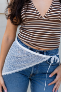 A close-up of a person wearing a brown and white striped crop top, blue jeans, and a white lace wrap tied around the waist.