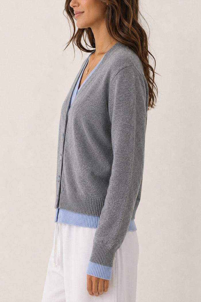 A woman wearing a gray knit cardigan with light blue ribbed trim at the V-neck, hem, and cuffs, paired with white pants against a plain light beige background.