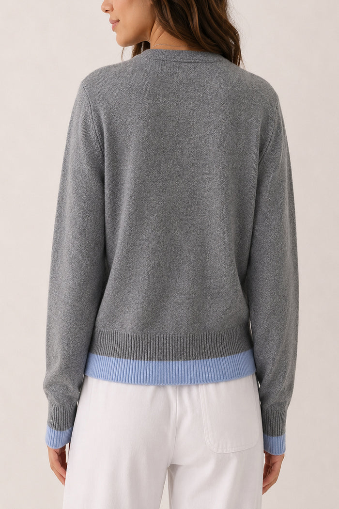 A woman shown from the back wearing a heather gray knit sweater with light blue ribbed trim at the hem and cuffs, paired with white pants, against a plain light beige studio background.