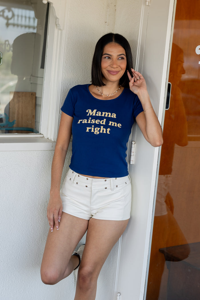Woman wearing a blue t-shirt with 'Mama raised me right' text, standing in front of a white door.