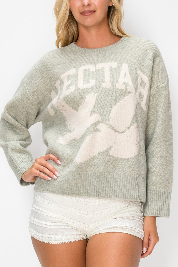 A woman wearing a light sage green knit sweater with a large cream 'NECTAR' text and dove graphic across the front, paired with white shorts.