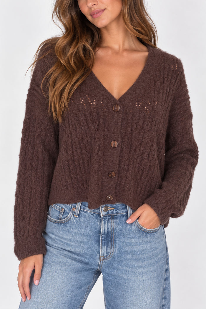 A woman wearing a brown cable-knit cardigan sweater with a V-neck and button-front closure, paired with light-wash blue jeans, against a plain light gray studio background.