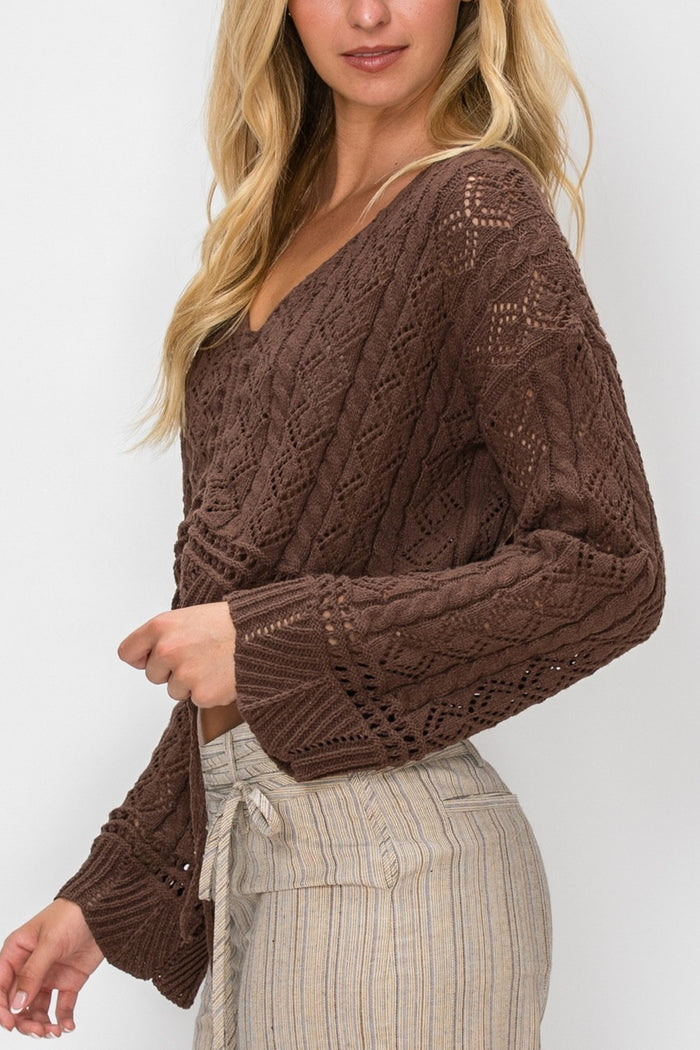 A woman wearing a brown cable-knit cropped sweater with a V-neck and long sleeves, paired with light beige striped pants, against a plain light gray background.