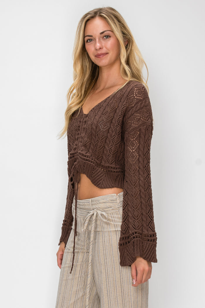A woman modeling a brown open-knit cropped sweater with long sleeves and a V-neck, paired with light beige drawstring pants, against a plain white studio background.