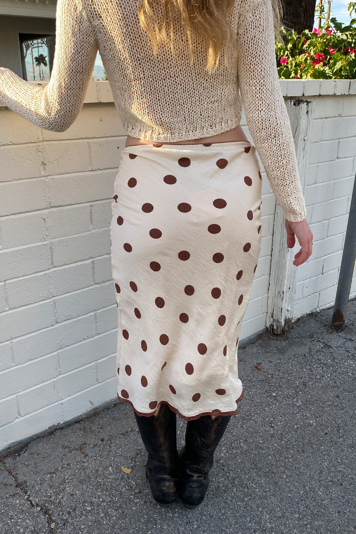 Person wearing a beige sweater and polka dot skirt against a white wall.