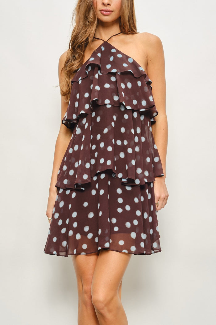 A woman wearing a brown halter-neck ruffled dress with white polka dots, shown from the waist up against a light gray studio background.