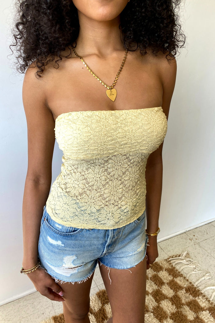 Woman wearing a yellow strapless top and denim shorts against a white wall.