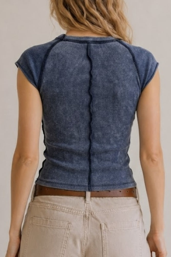 A woman viewed from the back wearing a dark blue ribbed short-sleeve crop top and light beige pants against a plain light background.