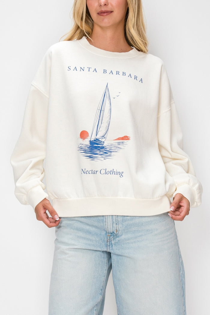 A woman wearing a cream-colored crewneck sweatshirt with a blue and orange sailboat graphic and the text 'SANTA BARBARA' and 'Nectar Clothing'. She is also wearing light-wash blue jeans against a plain white background.