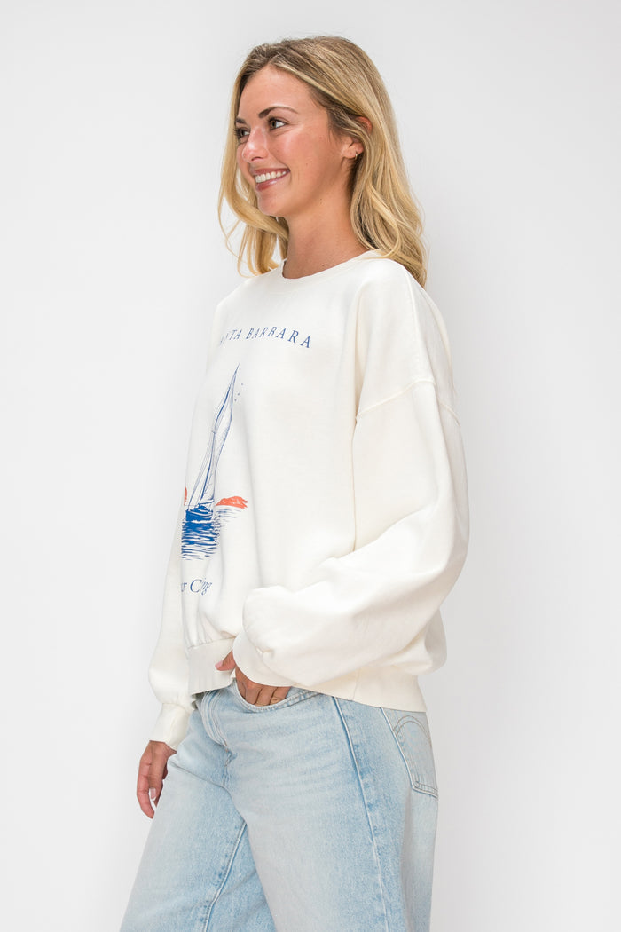 A woman wearing a cream-colored crewneck sweatshirt with a blue sailboat graphic and light-wash blue jeans, standing against a plain white studio background.