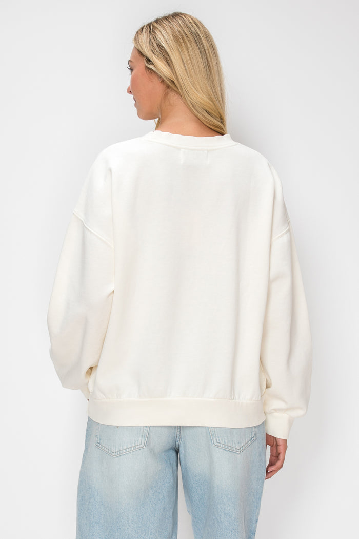 A woman viewed from the back wearing an oversized cream-colored crewneck sweatshirt and light blue jeans, set against a plain light gray studio background.