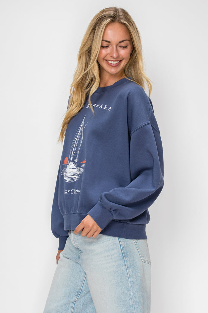 A woman wearing a navy blue crewneck sweatshirt with a sailboat graphic and the word 'Barbara' printed on the chest, paired with light-wash jeans, against a plain light gray background.