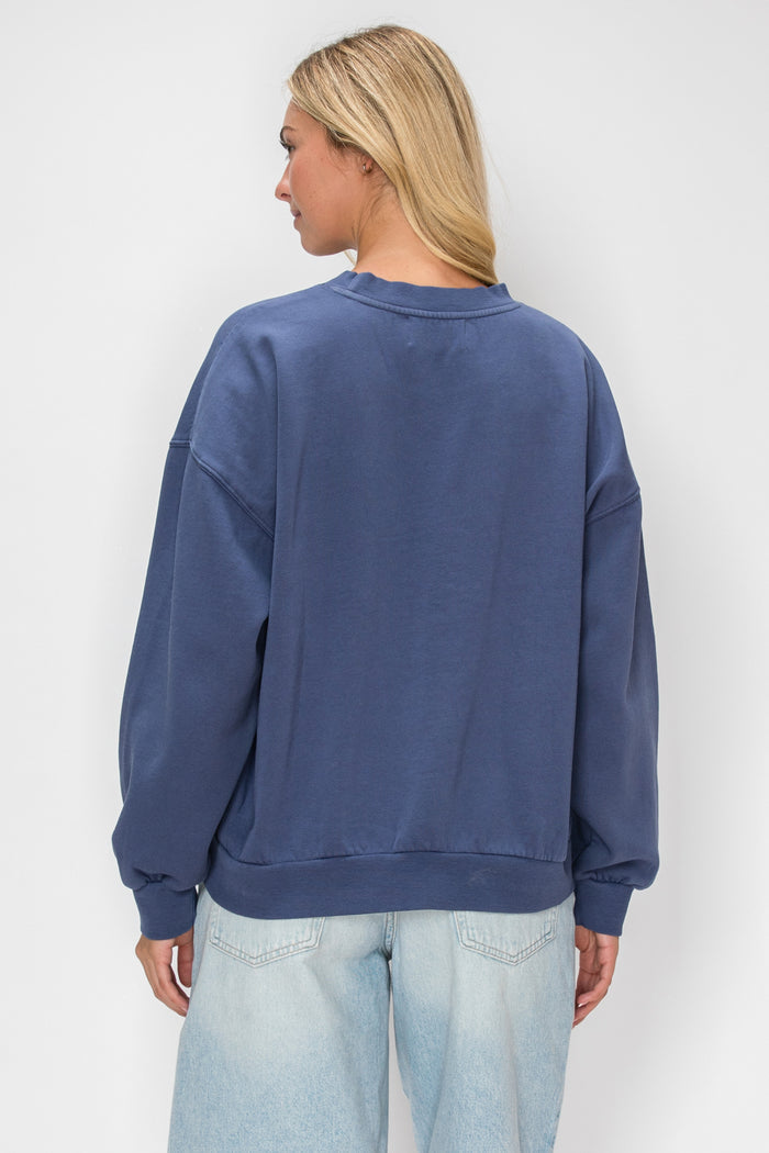 A woman viewed from the back wearing an oversized crewneck sweatshirt in a washed slate blue color, paired with light-wash jeans, against a plain light gray background.