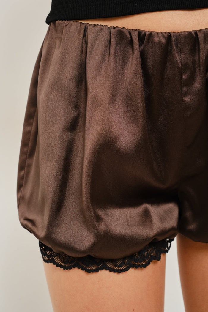 Close-up of a person wearing brown satin-like puff shorts with black lace trim at the hem, against a plain light background.