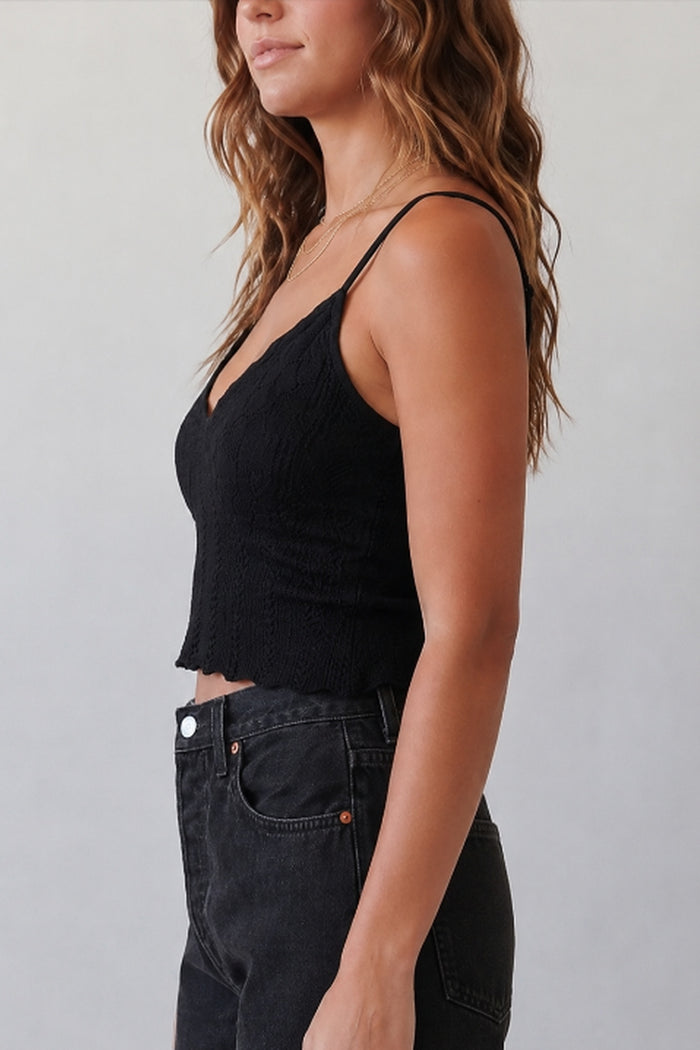 A woman shown in side profile wearing a black ribbed spaghetti-strap crop top and dark gray jeans against a plain light gray background.