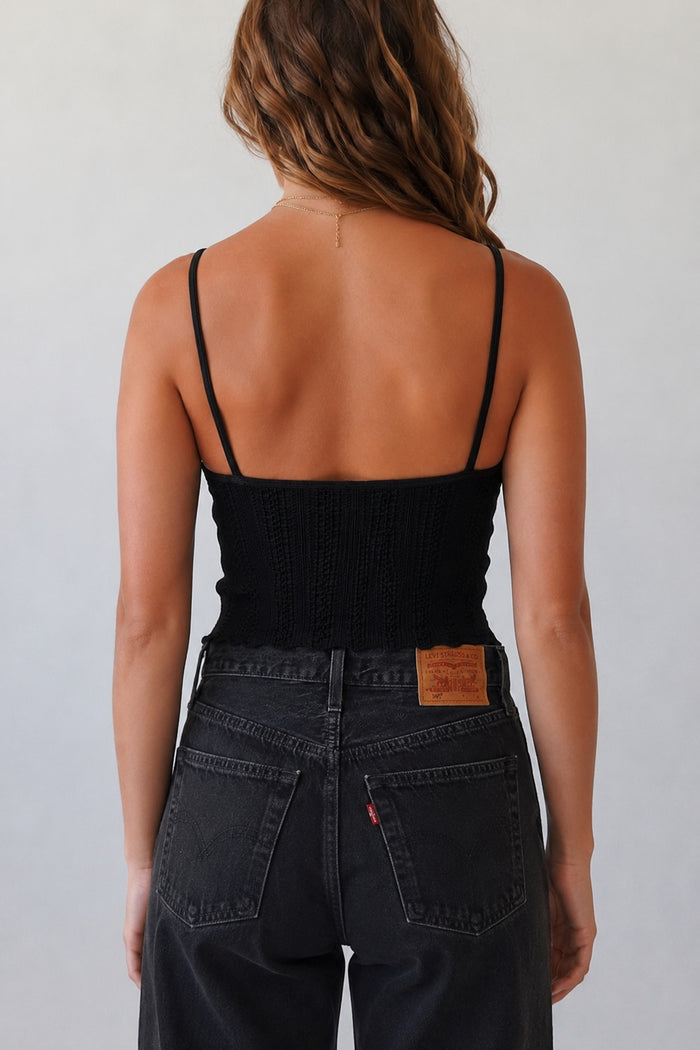 Rear view of a woman wearing a black ribbed spaghetti-strap camisole top and dark gray jeans against a light gray background.