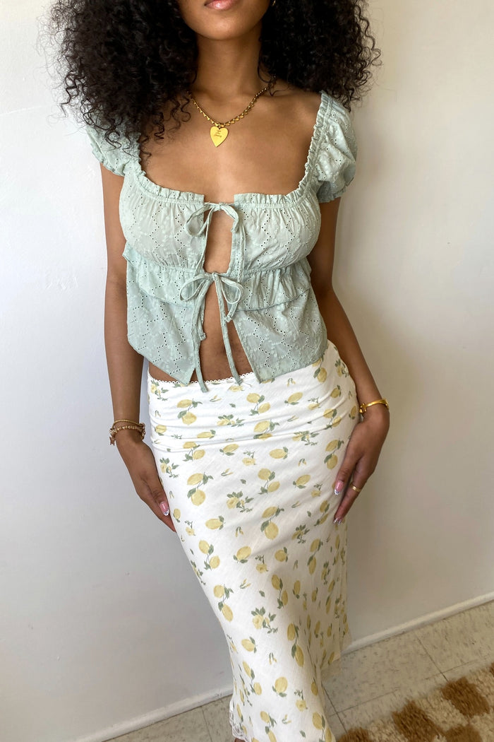 Woman wearing a green top with ruffled sleeves and a floral skirt against a plain background