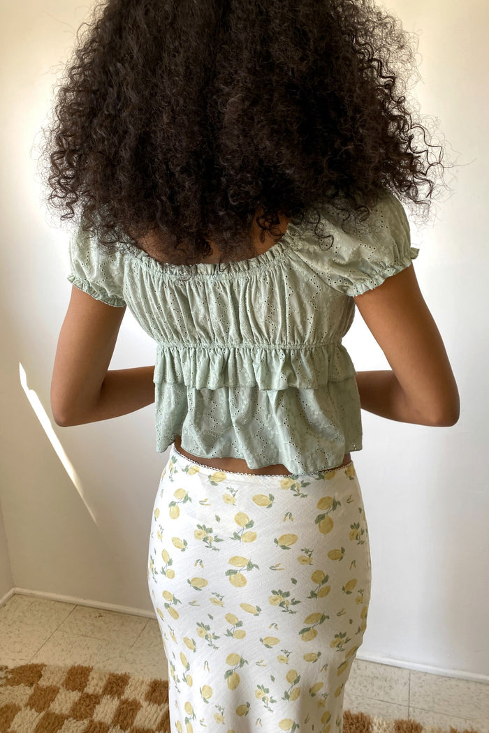 Woman wearing a green top with ruffled sleeves and a floral skirt against a plain background