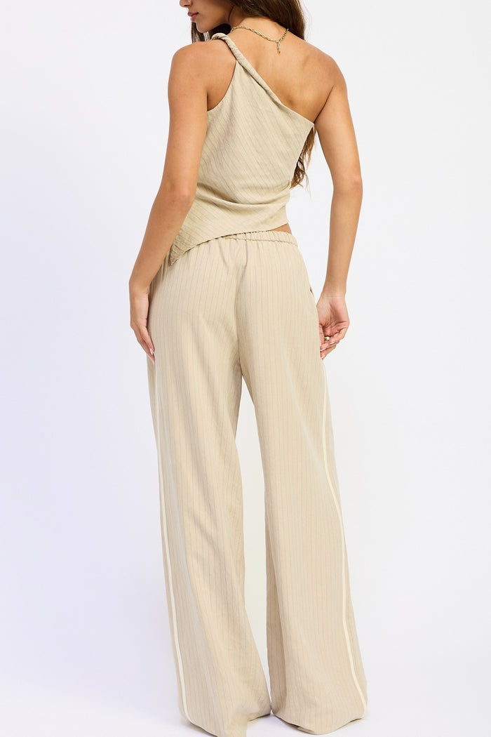A woman shown from the back wearing a beige ribbed two-piece set consisting of a one-shoulder top and matching wide-leg pants, against a plain white studio background.