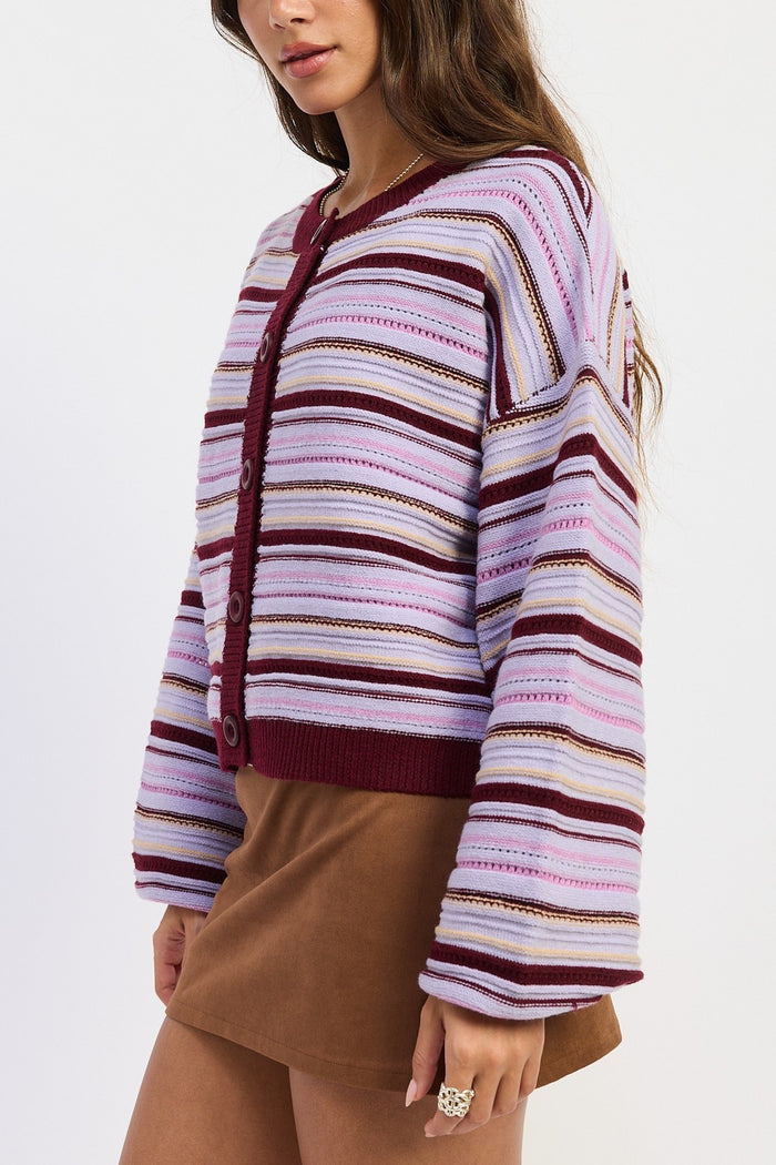 A woman wearing a cropped, long-sleeve knit cardigan with horizontal stripes in lavender, pink, burgundy, and beige, paired with brown shorts against a plain white background.