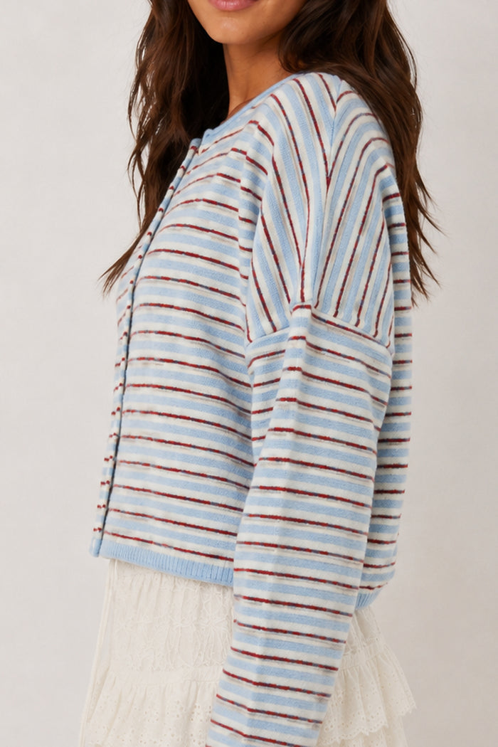 A woman wearing a cropped knit cardigan with horizontal light blue, white, and rust-brown stripes, paired with a white lace skirt against a plain light background.