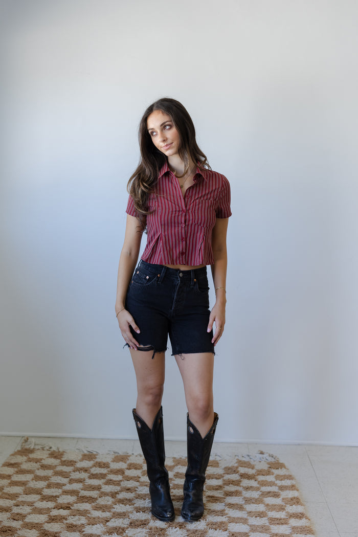 Woman wearing a red striped shirt, black shorts, and black boots standing against a white wall.