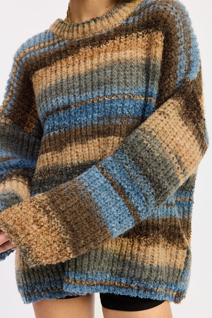 A close-up of a person wearing a chunky knit sweater with horizontal stripes in blue, tan, brown, and gray.